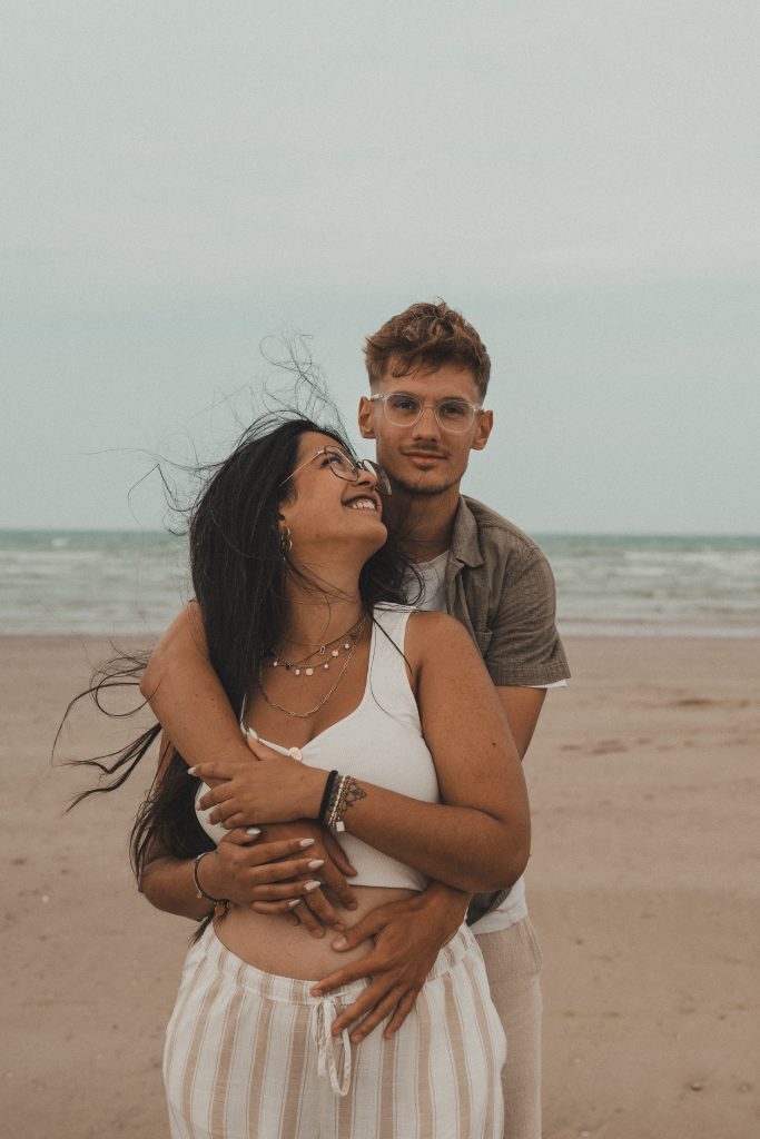 Couple sur la plage, femme souriante enlacée par son partenaire avec la mer en arrière-plan, séance photo