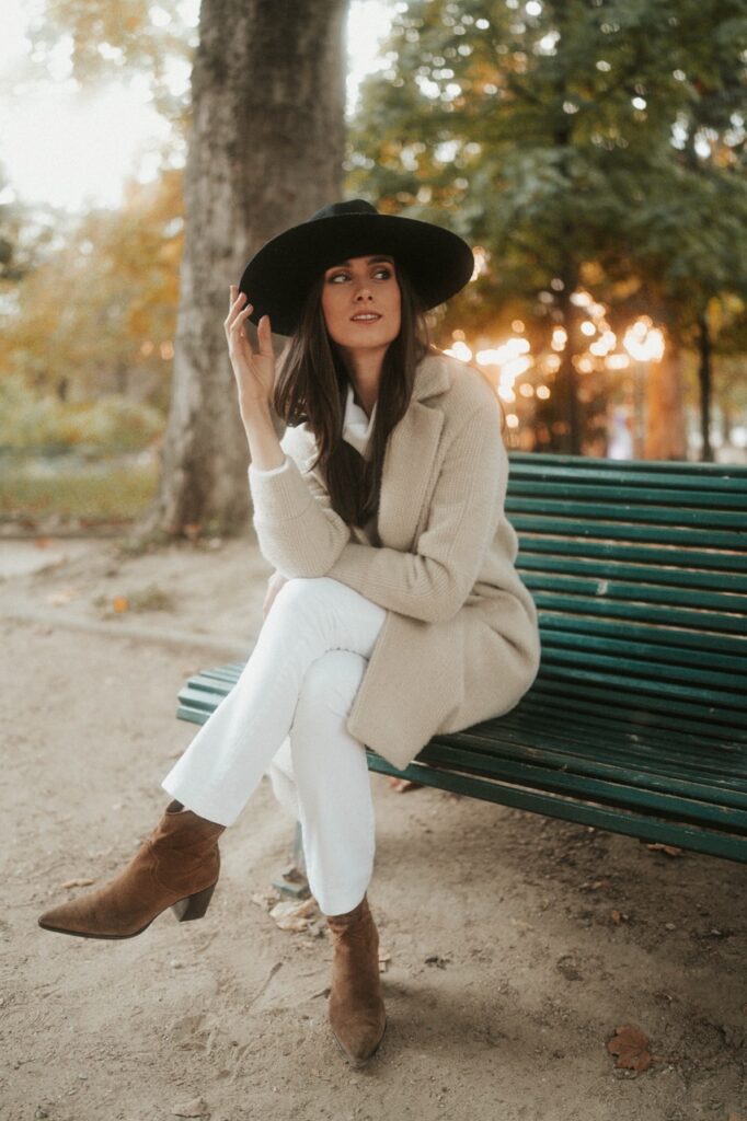 Femme en manteau beige et chapeau noir assise sur un banc de parc en automne, séance photo mode lifestyle